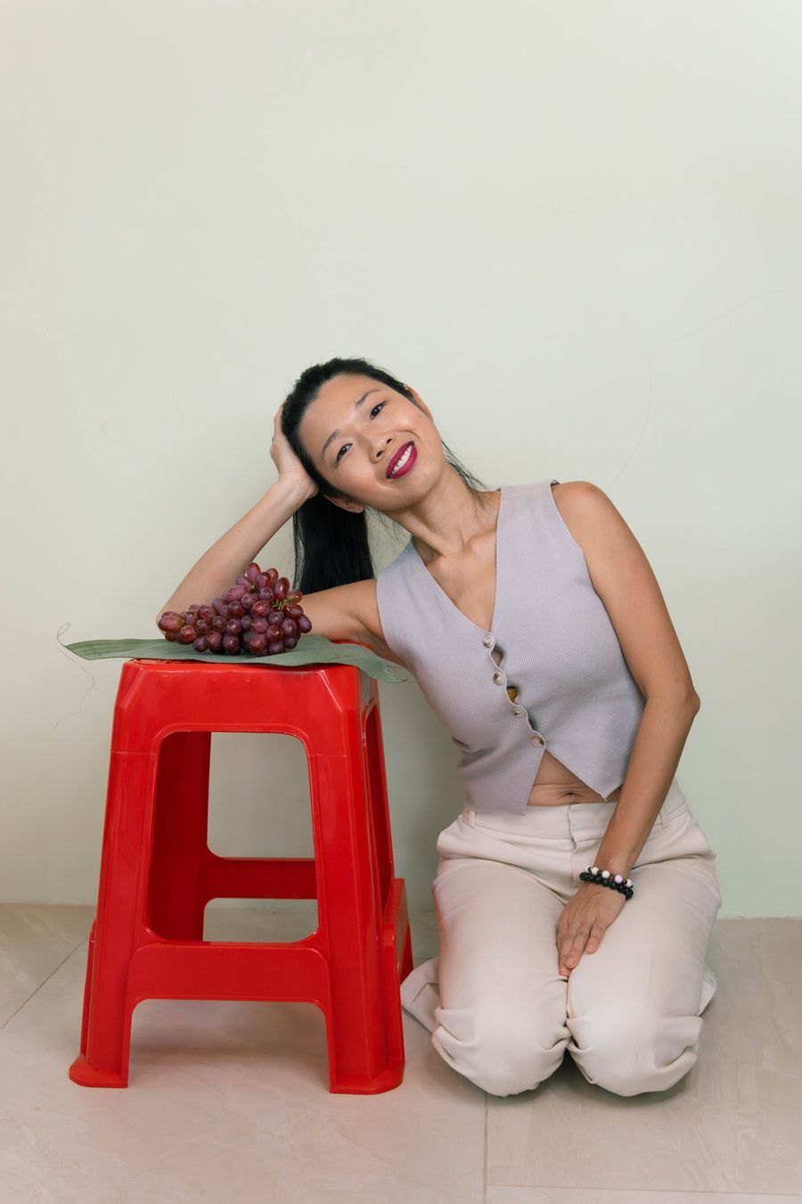 Tammie kneeling beside a red stool, resting her head on her arm next to a bunch of grapes