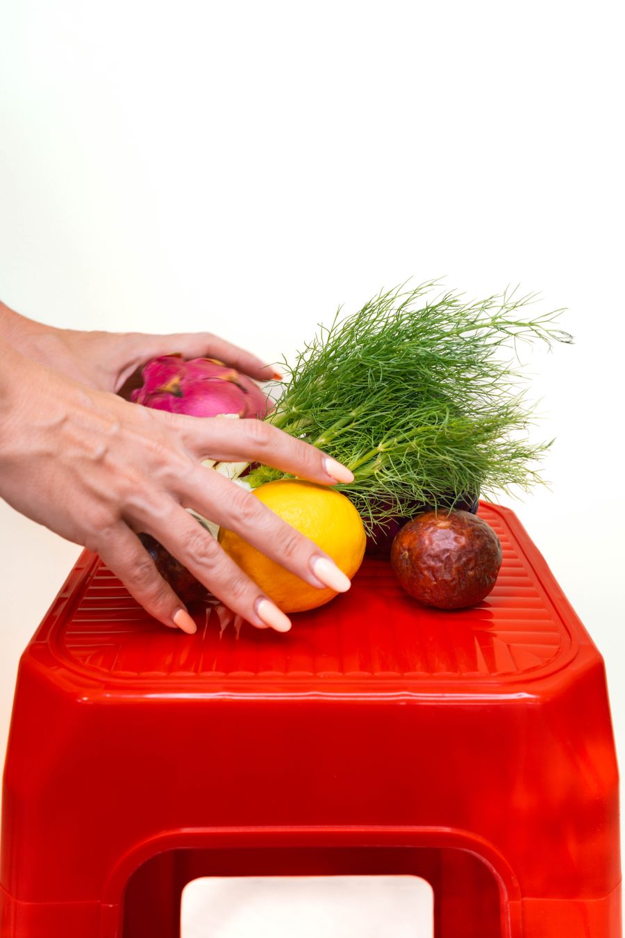 Hands arranging dragon fruit, fennel, a lemon and a passion fruit on a red stool